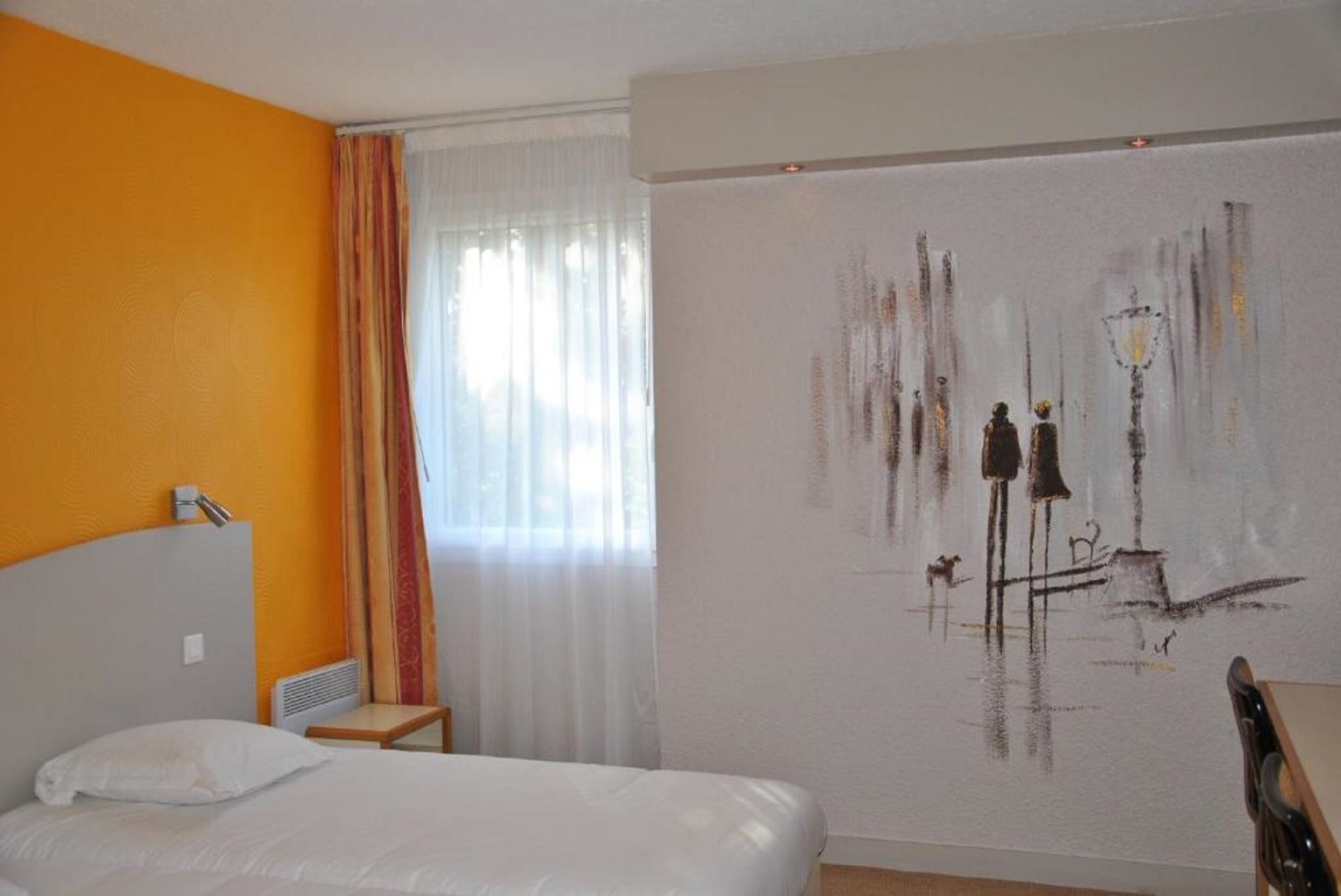 A hotel room with a bed, a small table, a chair, a window with curtains, and a wall with a painting.