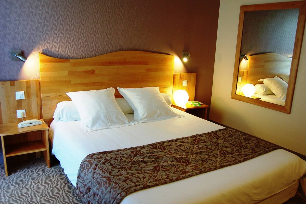 A hotel room with a wooden headboard, a bed with white linens, a bedside table with a lamp, and a mirror on the wall.