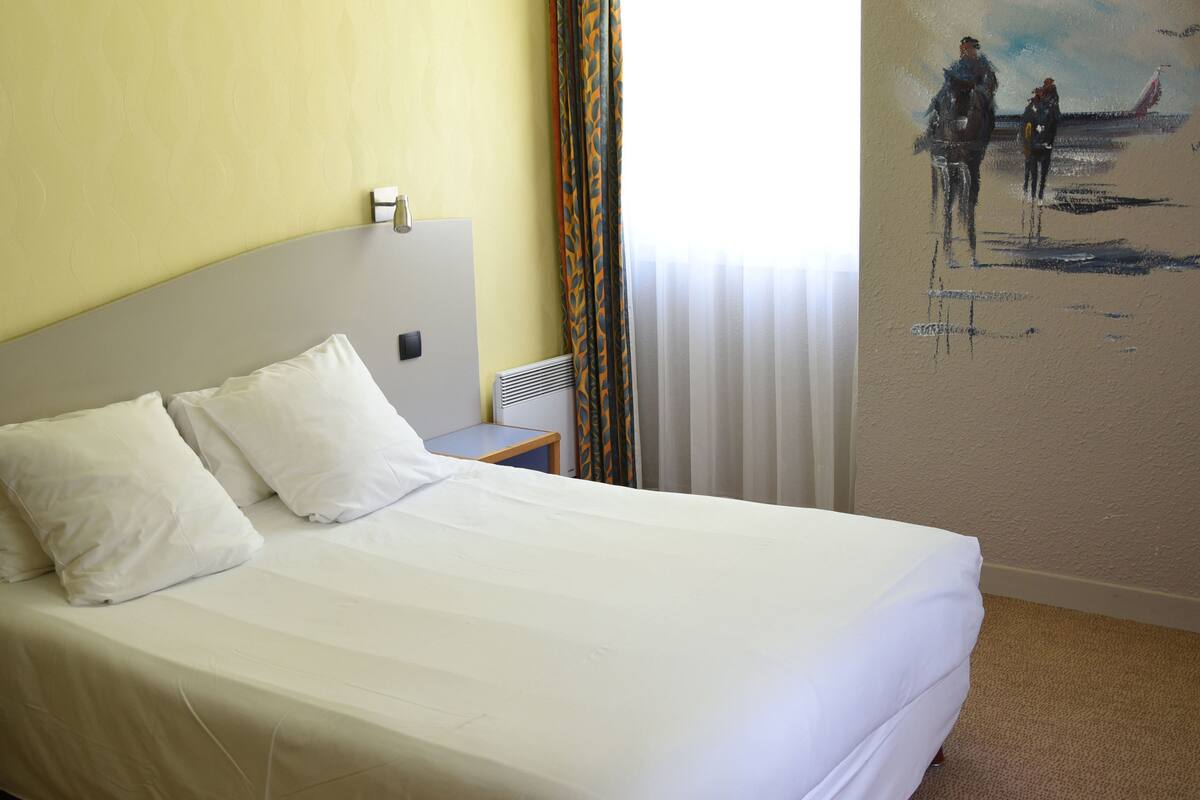 A hotel room with a bed, a nightstand, a lamp, a painting on the wall, and a window with curtains.