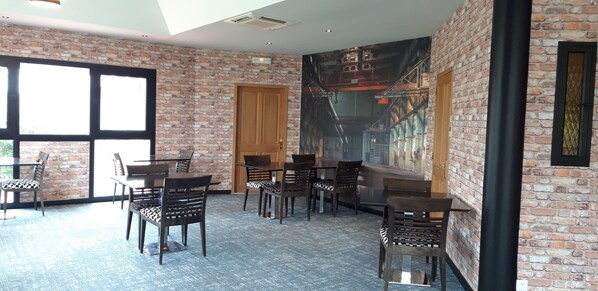 A dining area with brick walls, large windows, and a mural. Tables and chairs are arranged for dining. - Hôtel Cositel Coutances (Coutances)