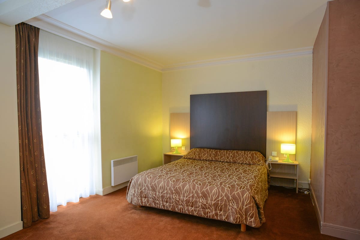 A hotel room with a large bed, two bedside lamps, a heater, and a window with curtains.