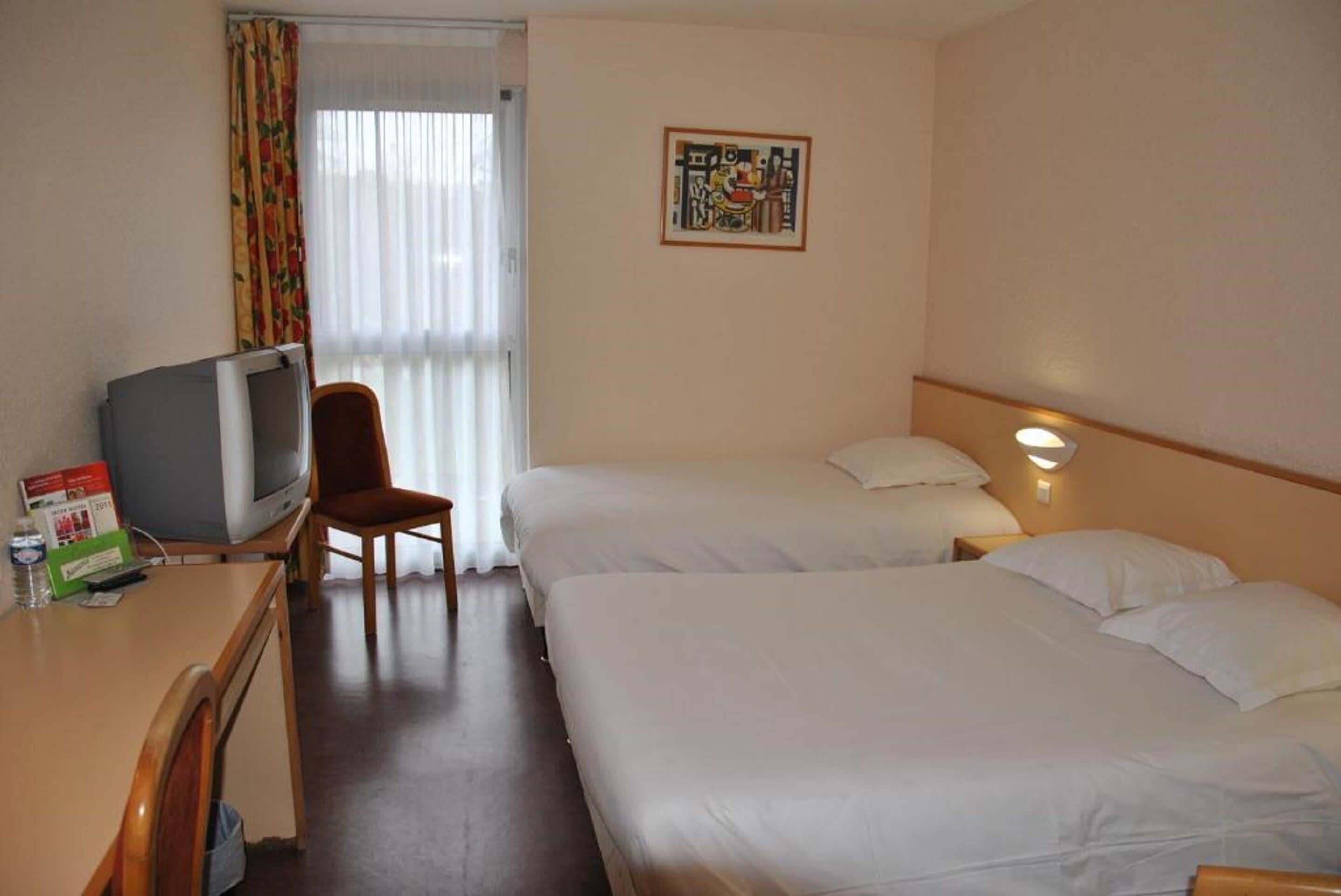 A hotel room with two beds, a desk with a television, a chair, and a window with curtains.