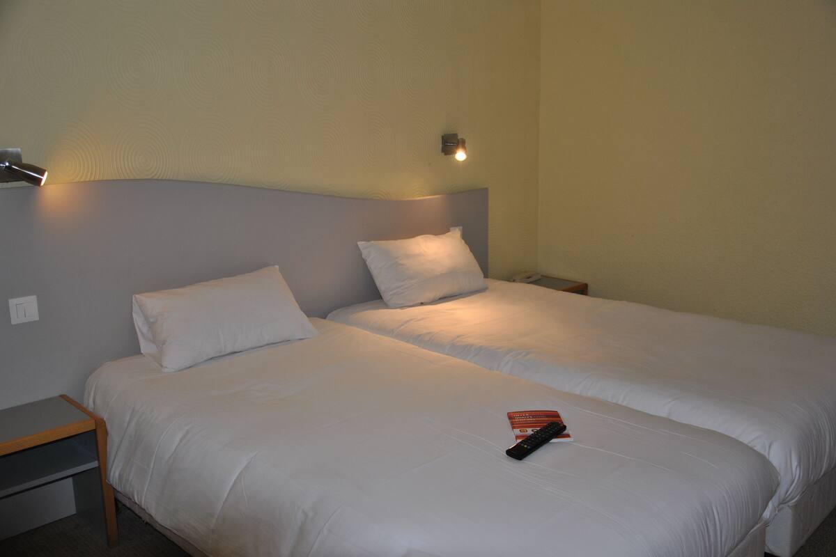 A double bed with white bedding, a bedside table, a nightstand, and a wall-mounted light fixture.