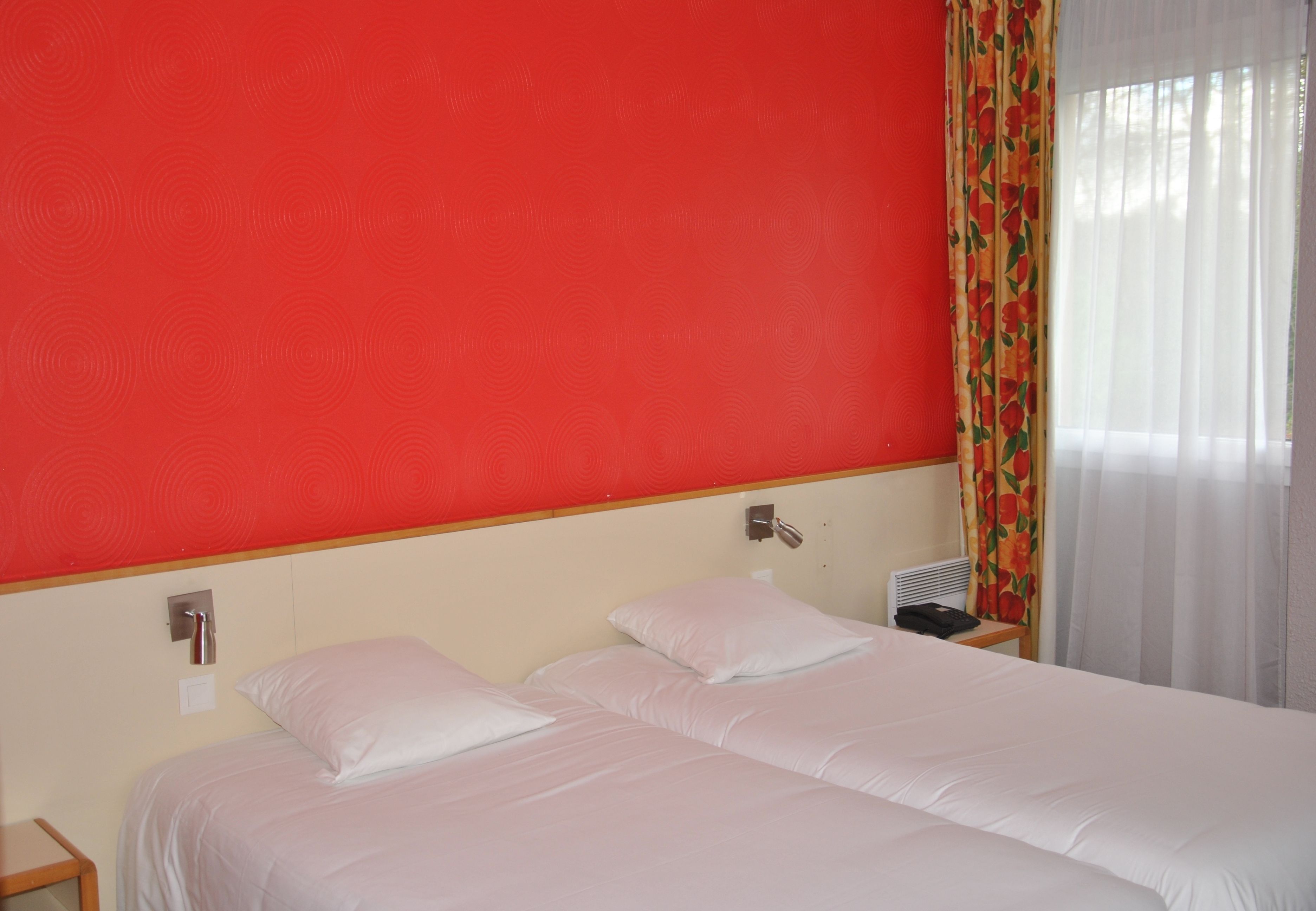 A hotel room with a red wall, two beds with white bedding, a bedside table with a lamp, and a window with sheer curtains.