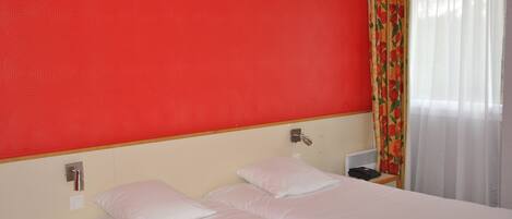 A hotel room with a red wall, two beds with white bedding, a bedside table with a lamp, and a window with sheer curtains.
