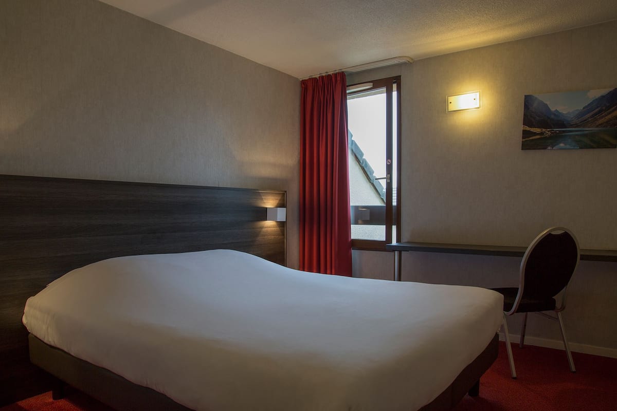 Habitación de hotel con una cama grande, un escritorio, una silla, una ventana con cortinas rojas y un cuadro en la pared.