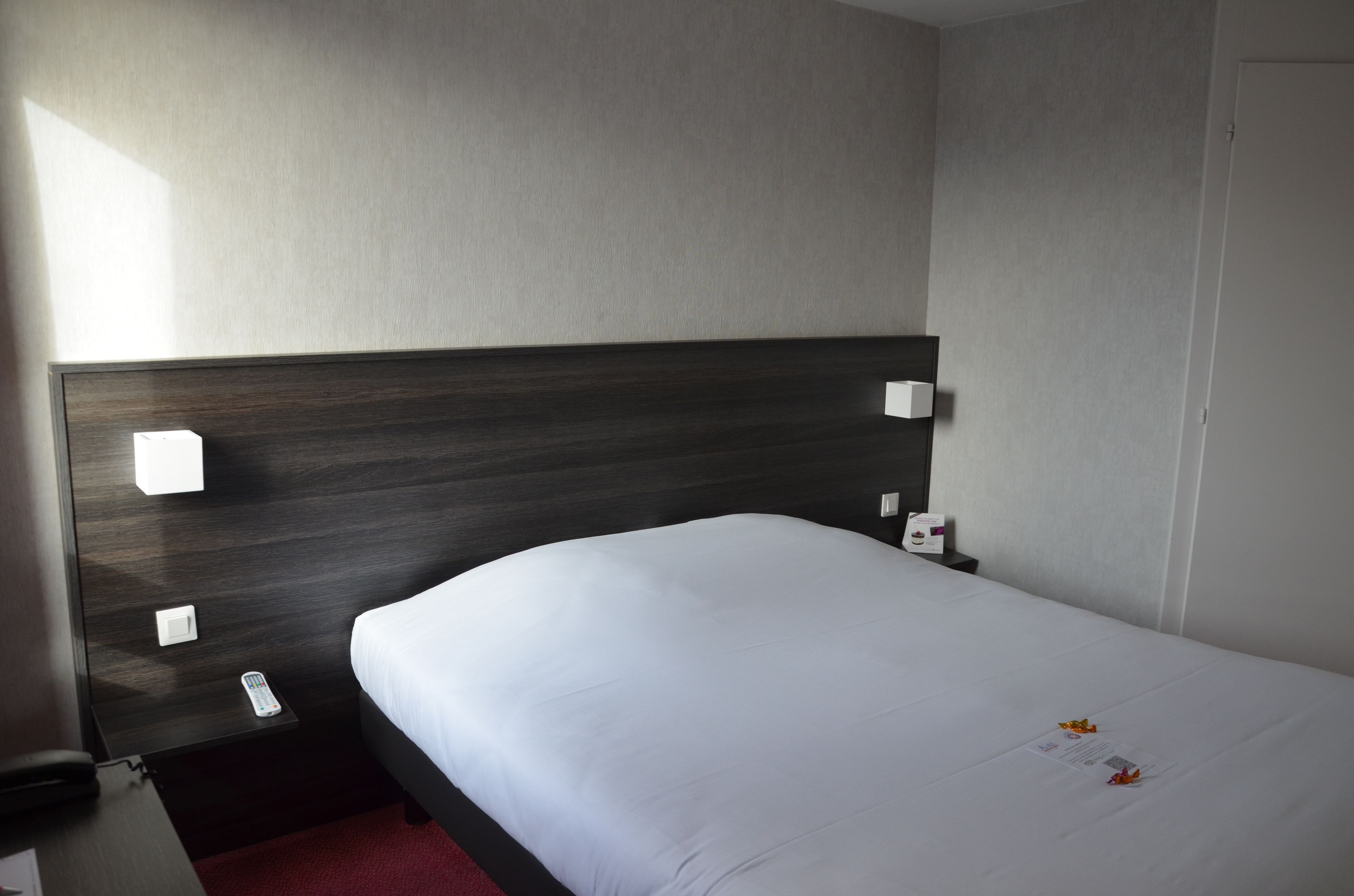 Habitación de hotel con tablero de cama de madera oscura, colcha blanca y control remoto sobre la mesita de noche.