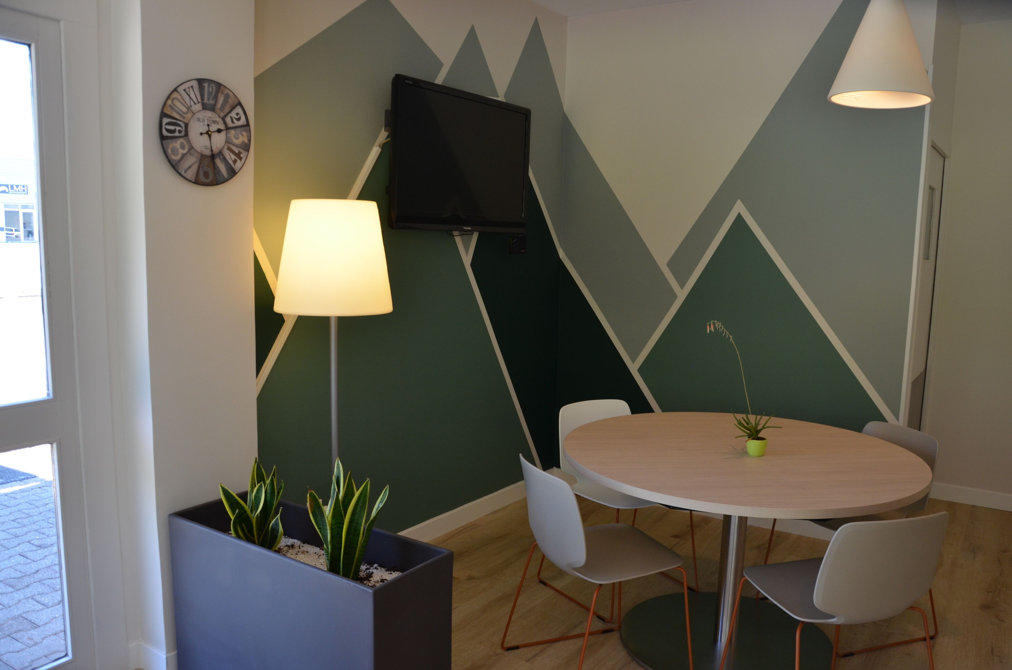 A conference room with a round table, white chairs, a TV mounted on the wall, a clock, and a lamp.