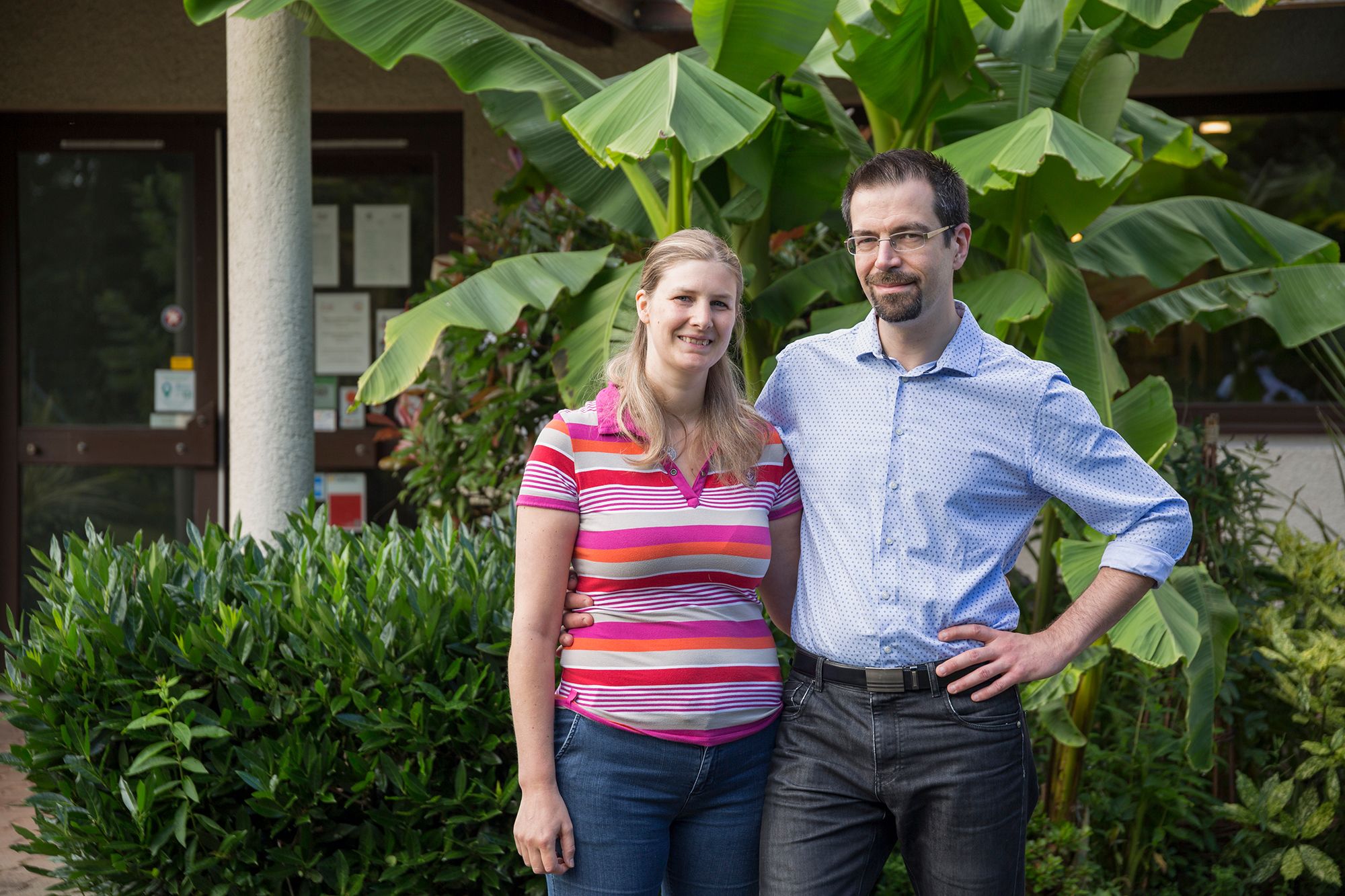 Ein Mann und eine Frau stehen vor einem Gebäude mit großen grünen Blättern und einer Glastür.