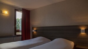 A hotel room with two beds, a window with red curtains, and wall-mounted lighting.