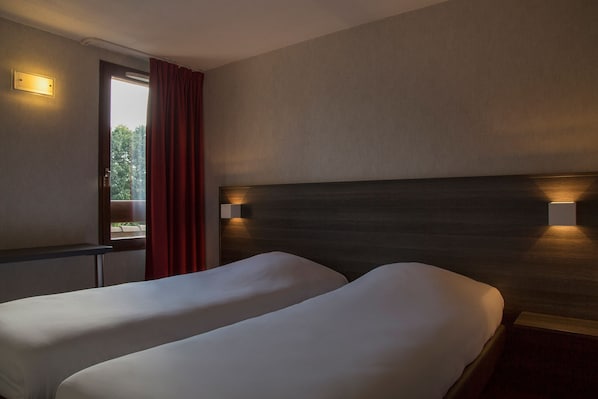 A hotel room with two beds, a window with red curtains, and wall-mounted lighting.