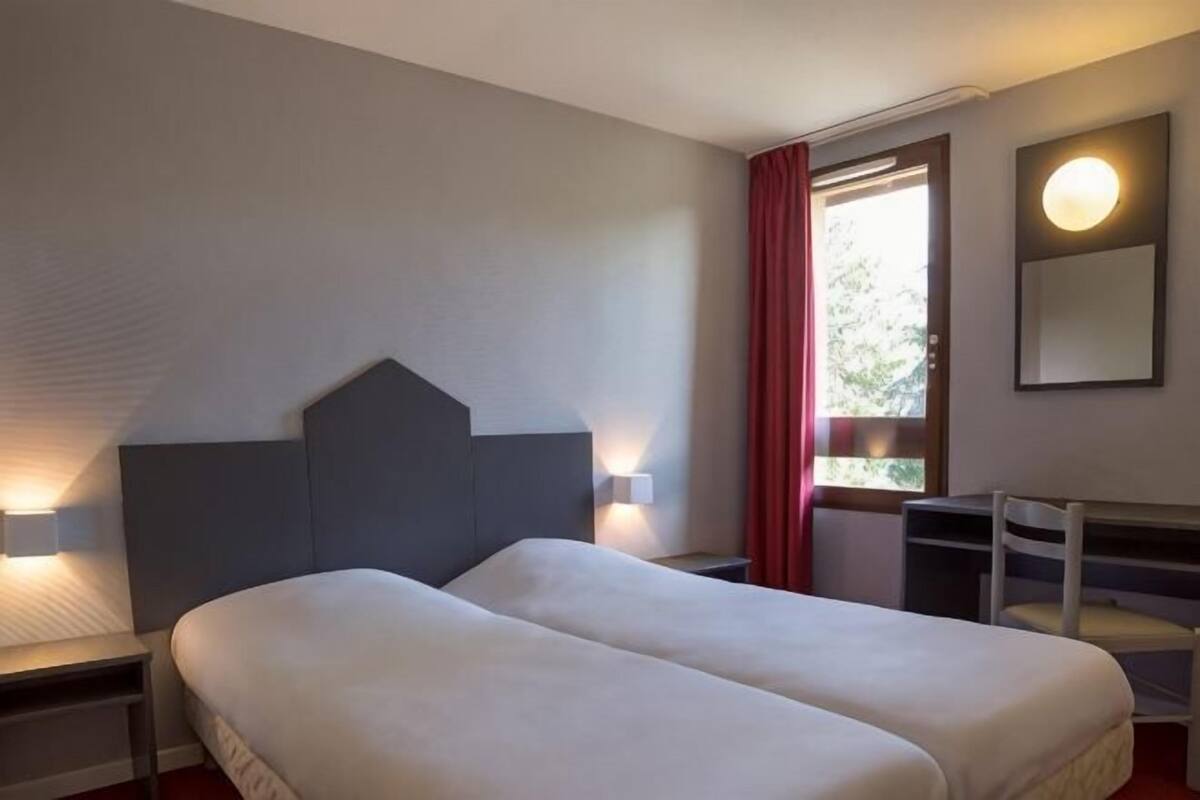 Een hotelkamer met twee bedden, een bureau, een stoel, een raam met gordijnen en wandlampen.