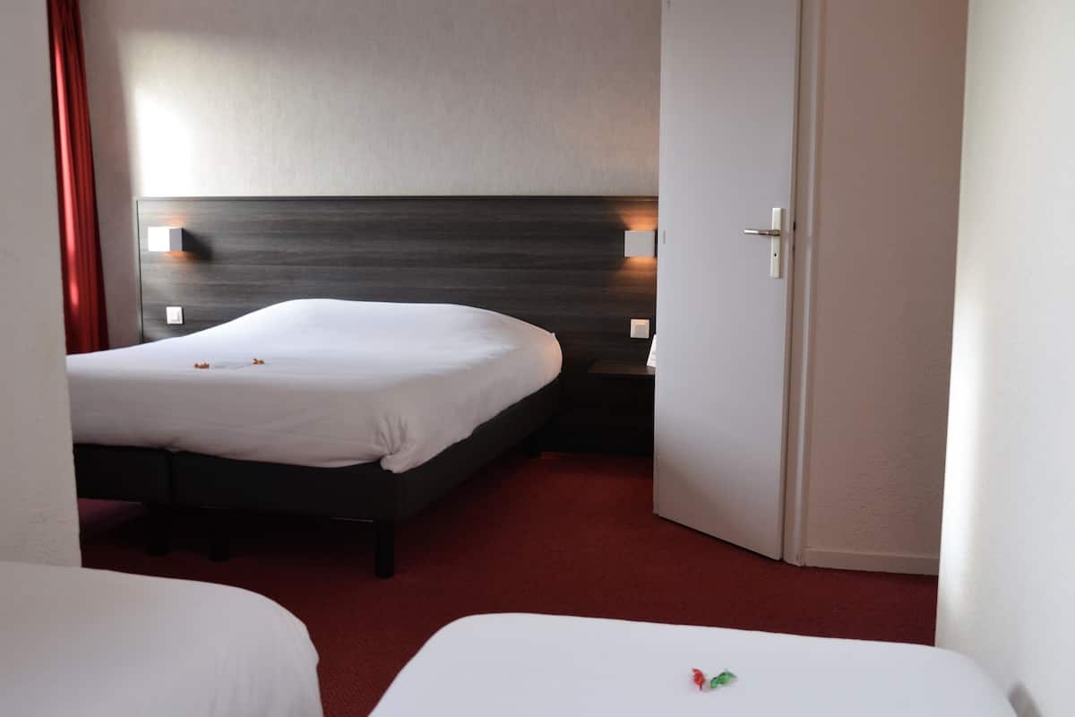 Habitación de hotel con dos camas individuales, cabecero de madera, alfombra roja y puerta blanca.