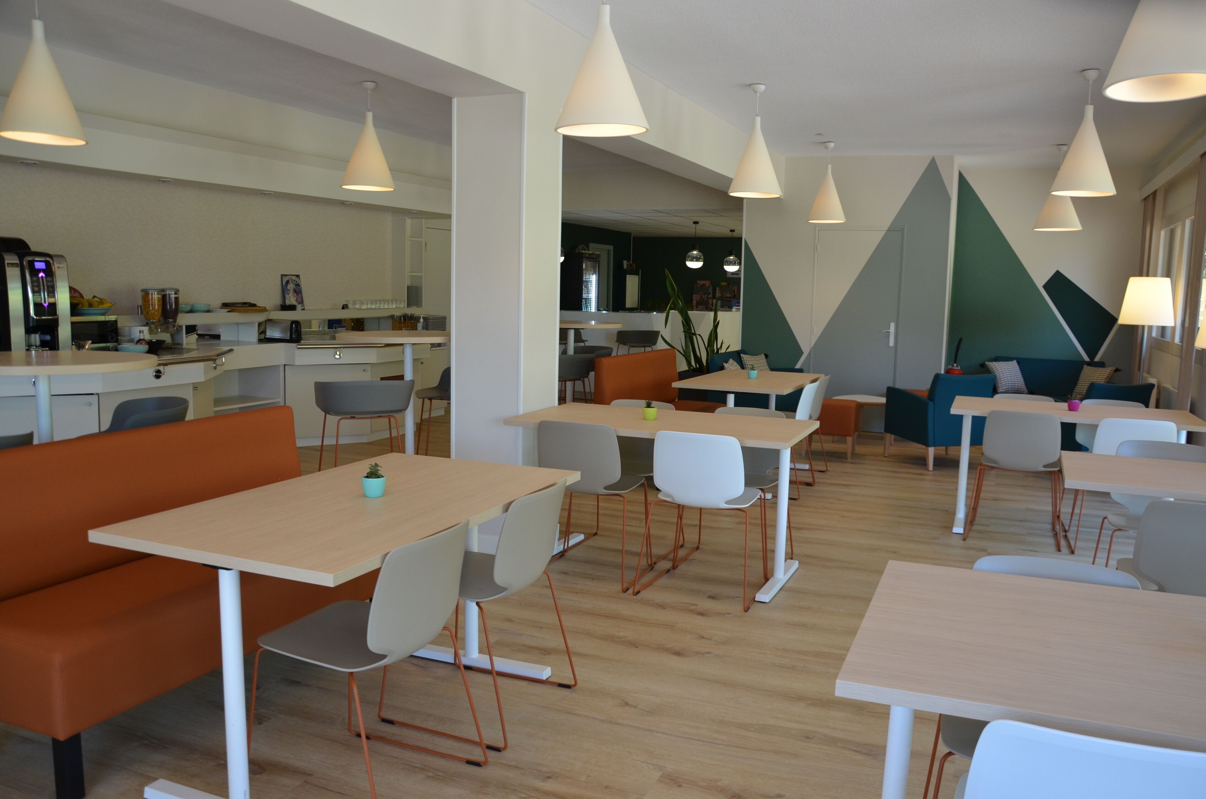 A modern, well-lit dining area with multiple tables and chairs, a kitchen in the background, and a seating area with a couch and chairs.