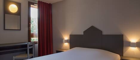 A hotel room with a bed, a desk, a chair, a mirror, and two wall-mounted lamps.