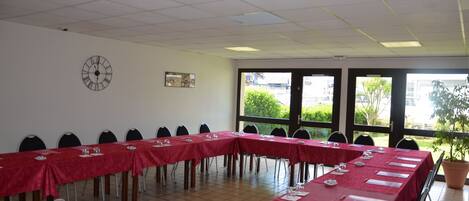 A conference room with long tables covered in red tablecloths, chairs, a clock on the wall, and large windows with a view of greenery outside.