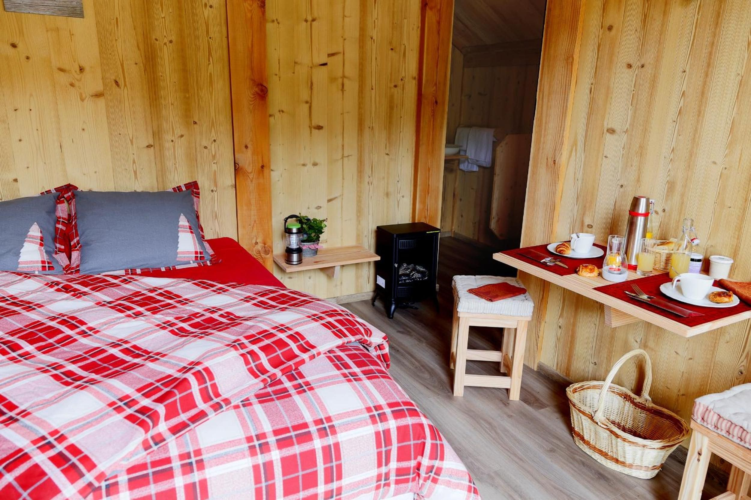 Una habitación de madera con una cama, una mesita con alimentos para el desayuno, una silla y una cesta.