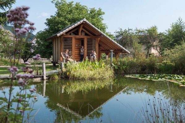 A wooden cabin with a sloped roof, situated by a calm pond with lily pads, surrounded by lush greenery and trees. - Logis Hotel la Résidence (Le Val-d'Ajol)