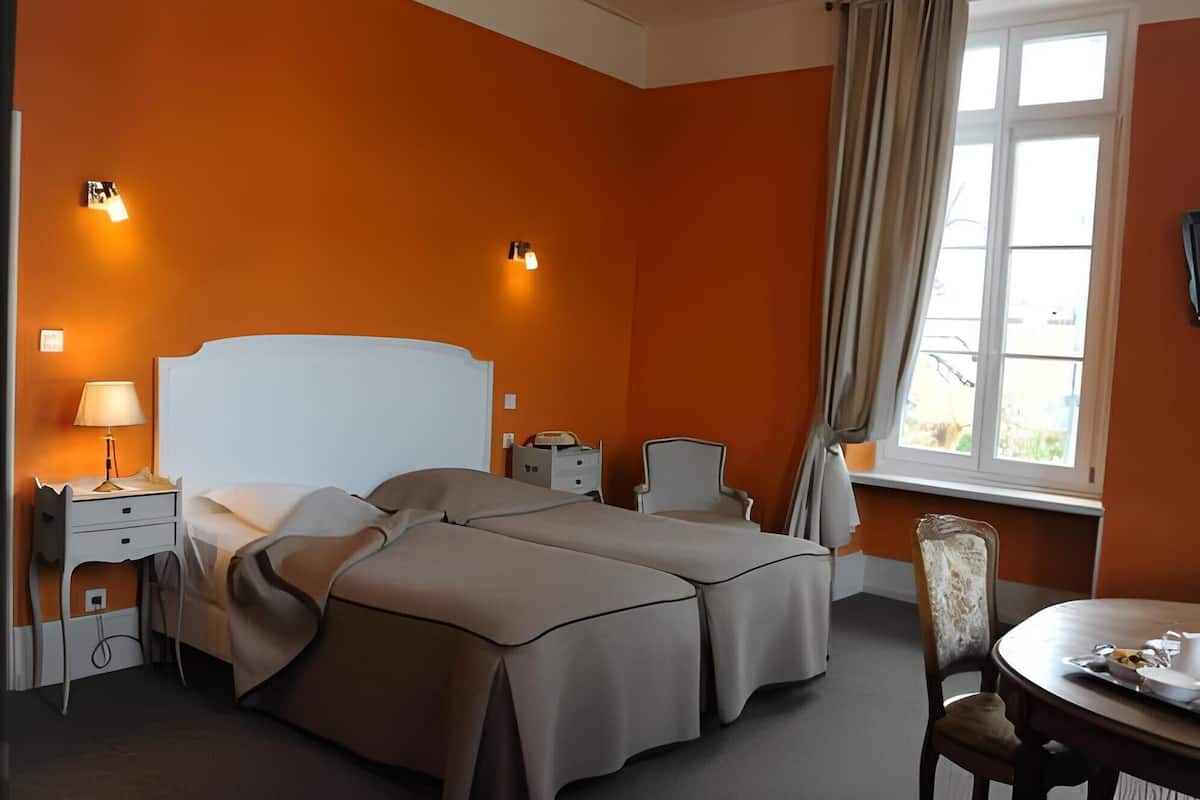 A hotel room with two beds, a desk with a lamp, a chair, a dining table with a tray, and a window with curtains.