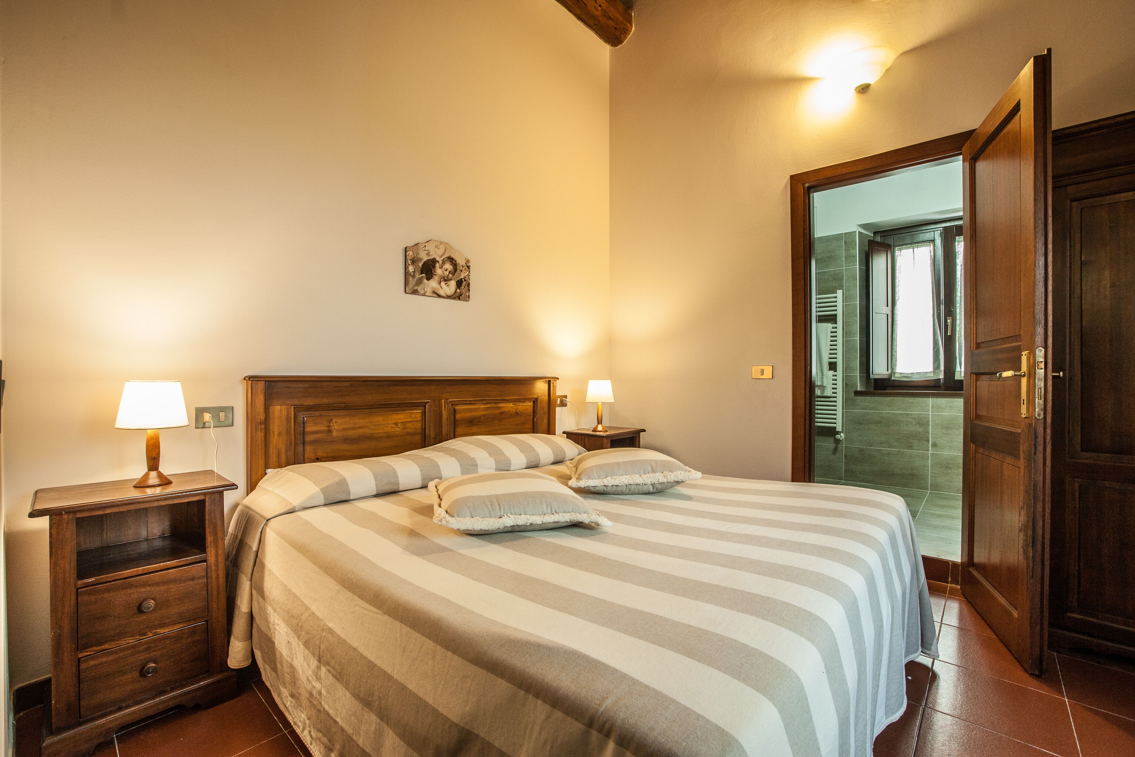 Una cama bien hecha con ropa de cama a rayas y dos almohadas, una mesita de noche de madera con una lámpara y una puerta que da a un baño.