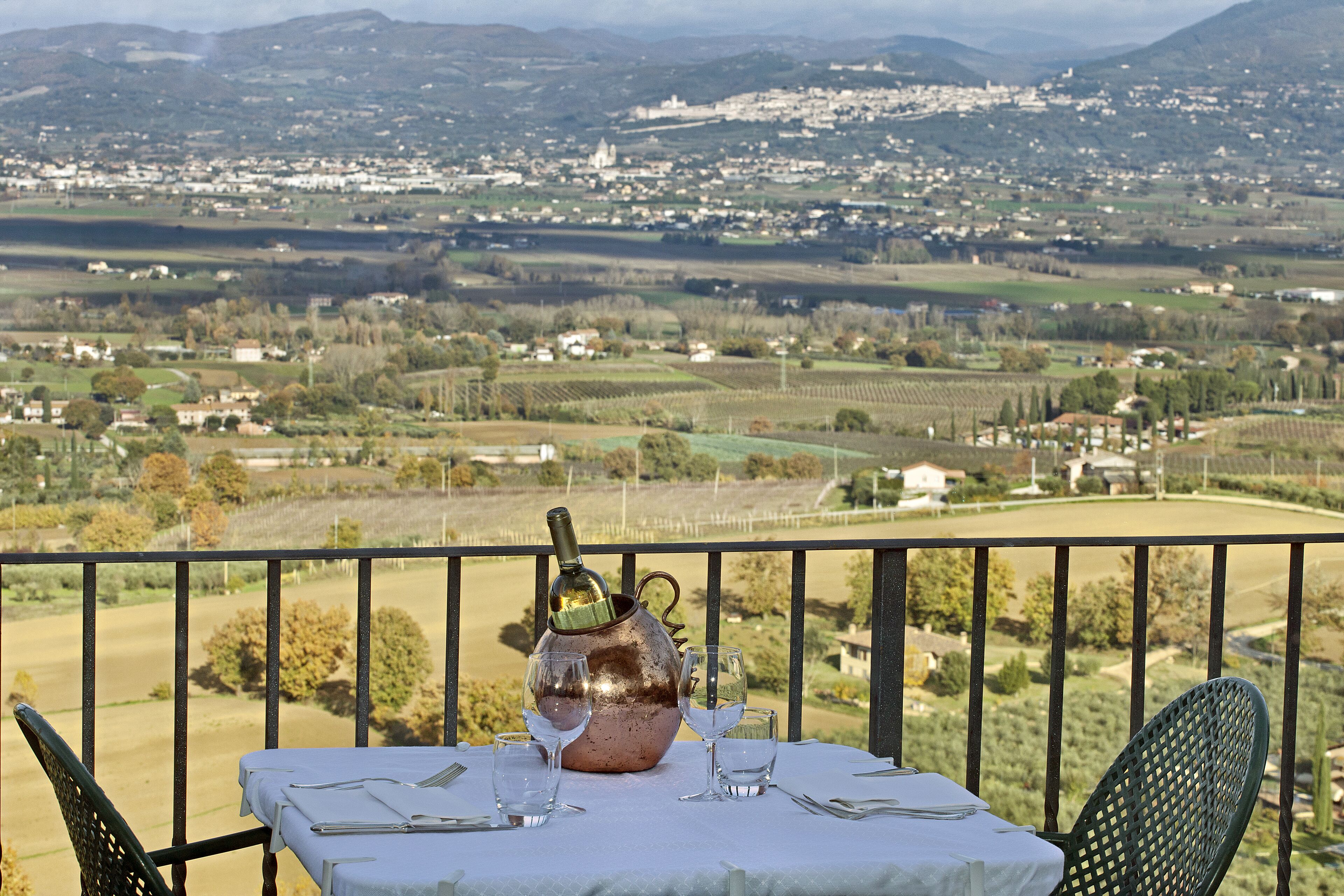 A table set for two with a copper jug and wine glasses on a balcony overlooking a rural landscape.
