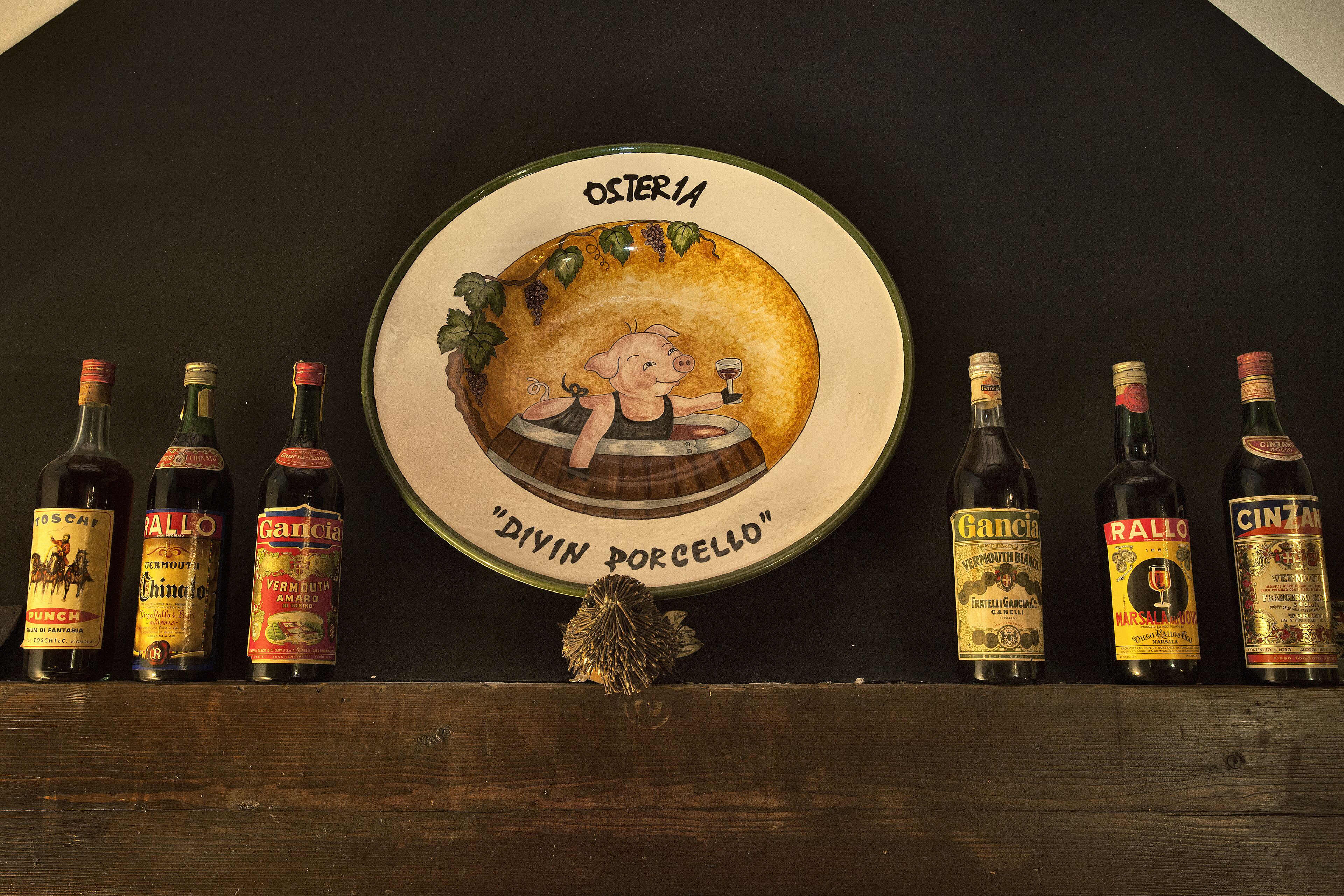 A collection of wine bottles with labels such as "PERSCH", "Rallo", and "Gancia" on a wooden shelf.