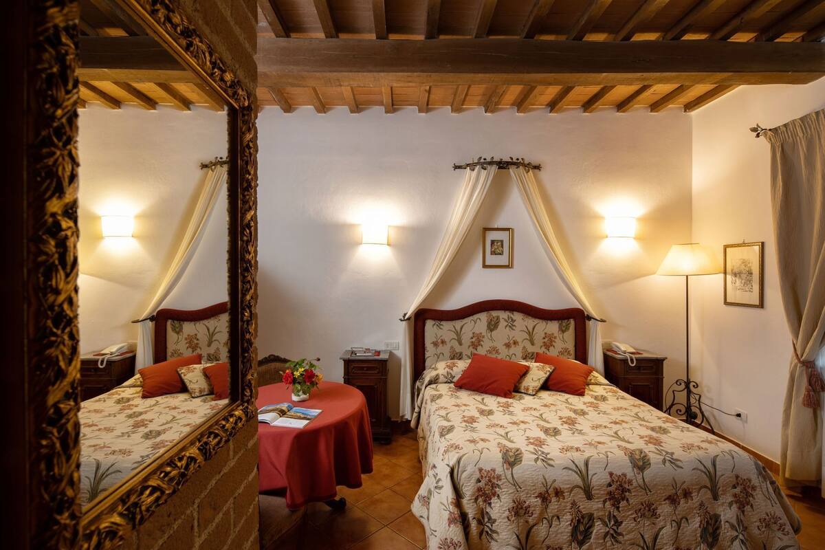 Une chambre avec deux lits, une table recouverte d’une nappe rouge, un miroir et des lampes fixées au mur.