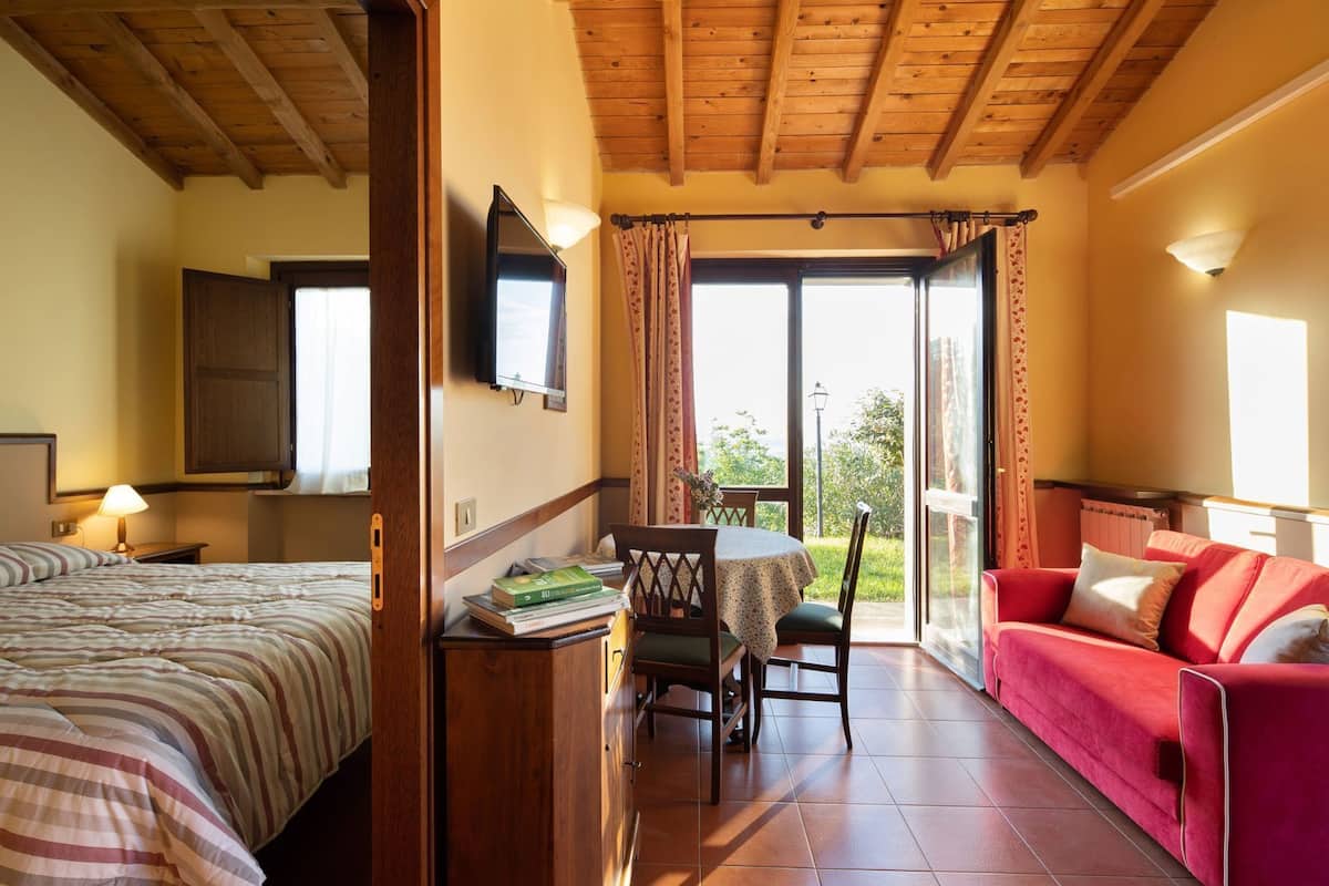 Une chambre d’hôtel avec un lit, un bureau avec une chaise, une table avec une chaise, un canapé rouge et une grande fenêtre.