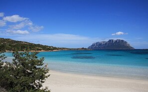 A beach with clear turquoise waters, a sandy shore, and a prominent rocky mountain in the background. - Hotel Daniel (Olbia)