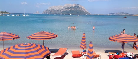 Une plage avec des parasols à rayures, des chaises longues et une vue dégagée sur la mer et les montagnes lointaines.