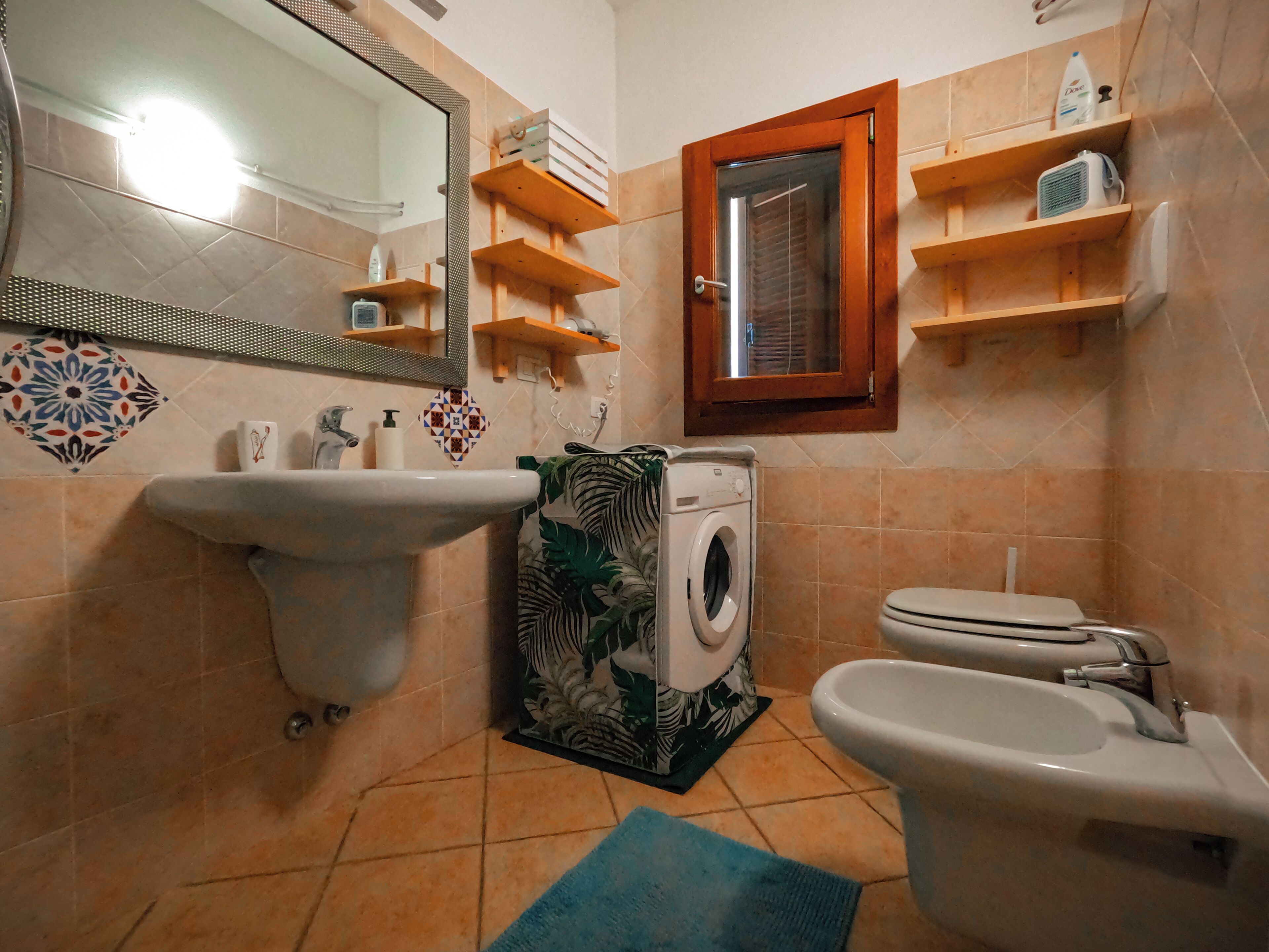 A bathroom with a sink, toilet, washing machine, and shelves with toiletries.