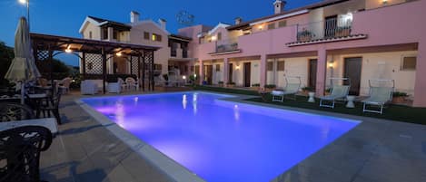 A swimming pool with a blue light, outdoor seating, and a building with balconies.