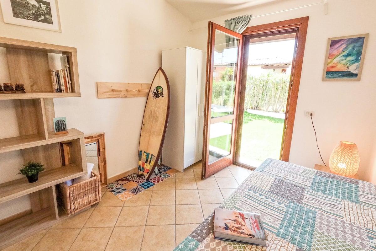 A room with a surfboard leaning against a wall, a bed with a patterned bedspread, a bookshelf with books and a cactus, and a door leading outside.