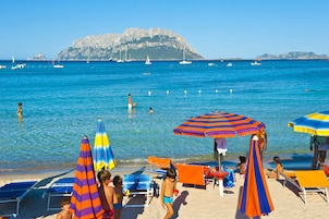 A beach with clear blue water, boats, and rocky cliffs in the background.