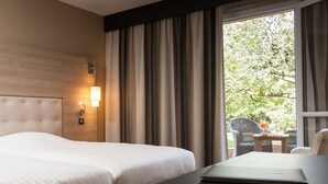 A hotel room with a large bed, a desk with a laptop, a nightstand, and a window with curtains.