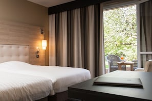 A hotel room with a large bed, a desk with a laptop, a nightstand, and a window with curtains.