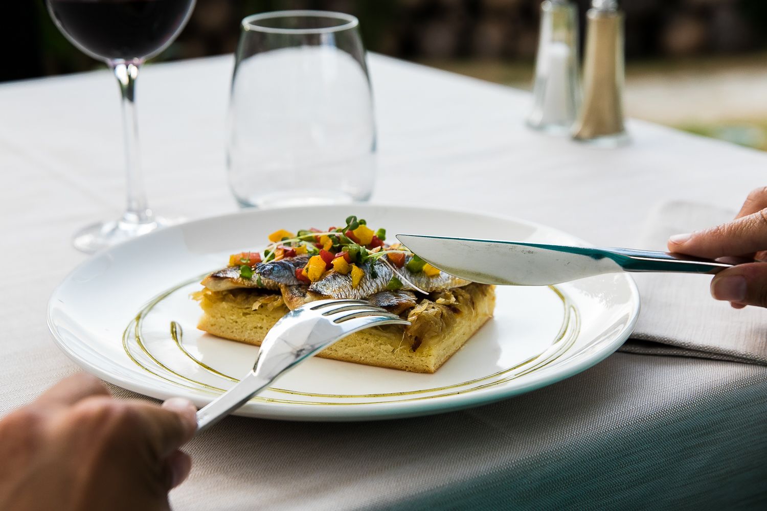 Un plato con un sándwich cubierto de pescado y verduras, un tenedor y un cuchillo sobre una mesa, con una copa de vino y salero al fondo.