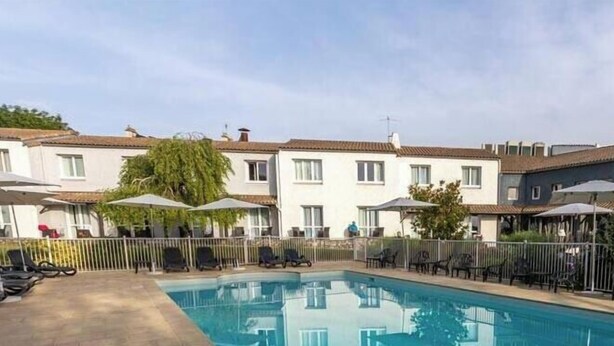 A swimming pool surrounded by lounge chairs and umbrellas, with a row of white buildings in the background.