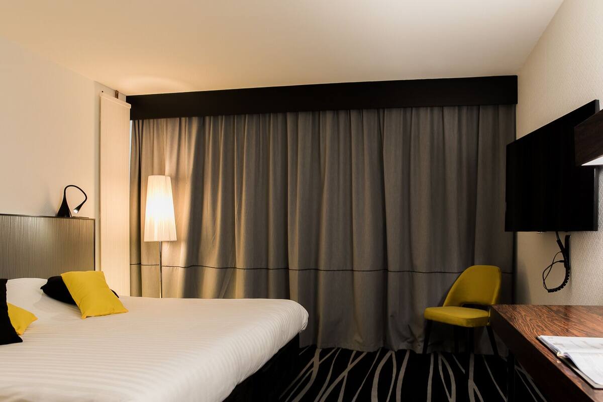 Una habitación de hotel con cama, escritorio, silla, televisor y ventana con cortinas.