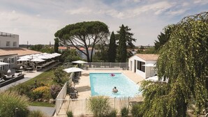 A pool area with a person swimming, surrounded by outdoor seating and umbrellas.