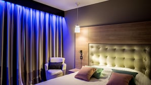 A modern bedroom with a tufted headboard, a bedside table, a chair, and a large window with curtains. - LOGIS HOTEL LE RABELAIS (Fontenay-le-Comte)