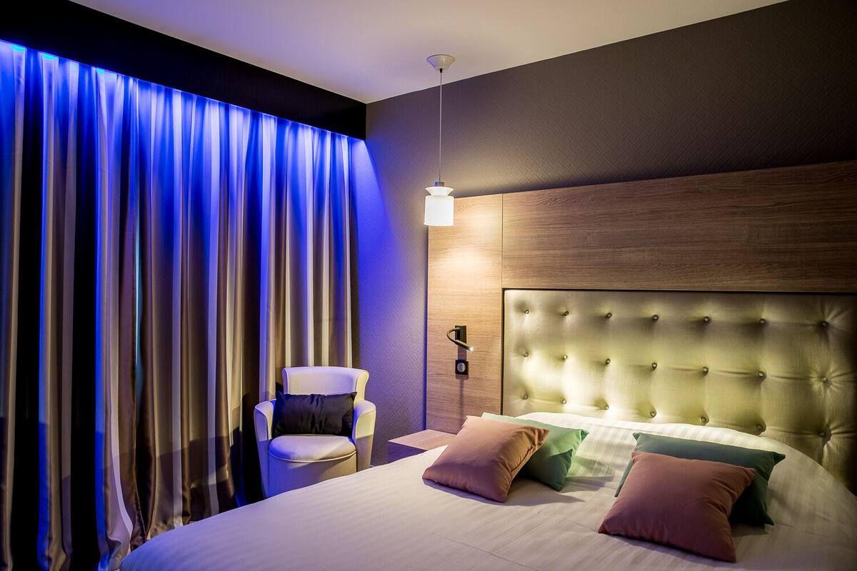 Un dormitorio moderno con un cabecero capitonado, una mesita de noche, una silla y un ventanal con cortinas.