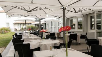 Een terras met tafels en stoelen onder witte parasols, omringd door een gebouw met grote ramen.