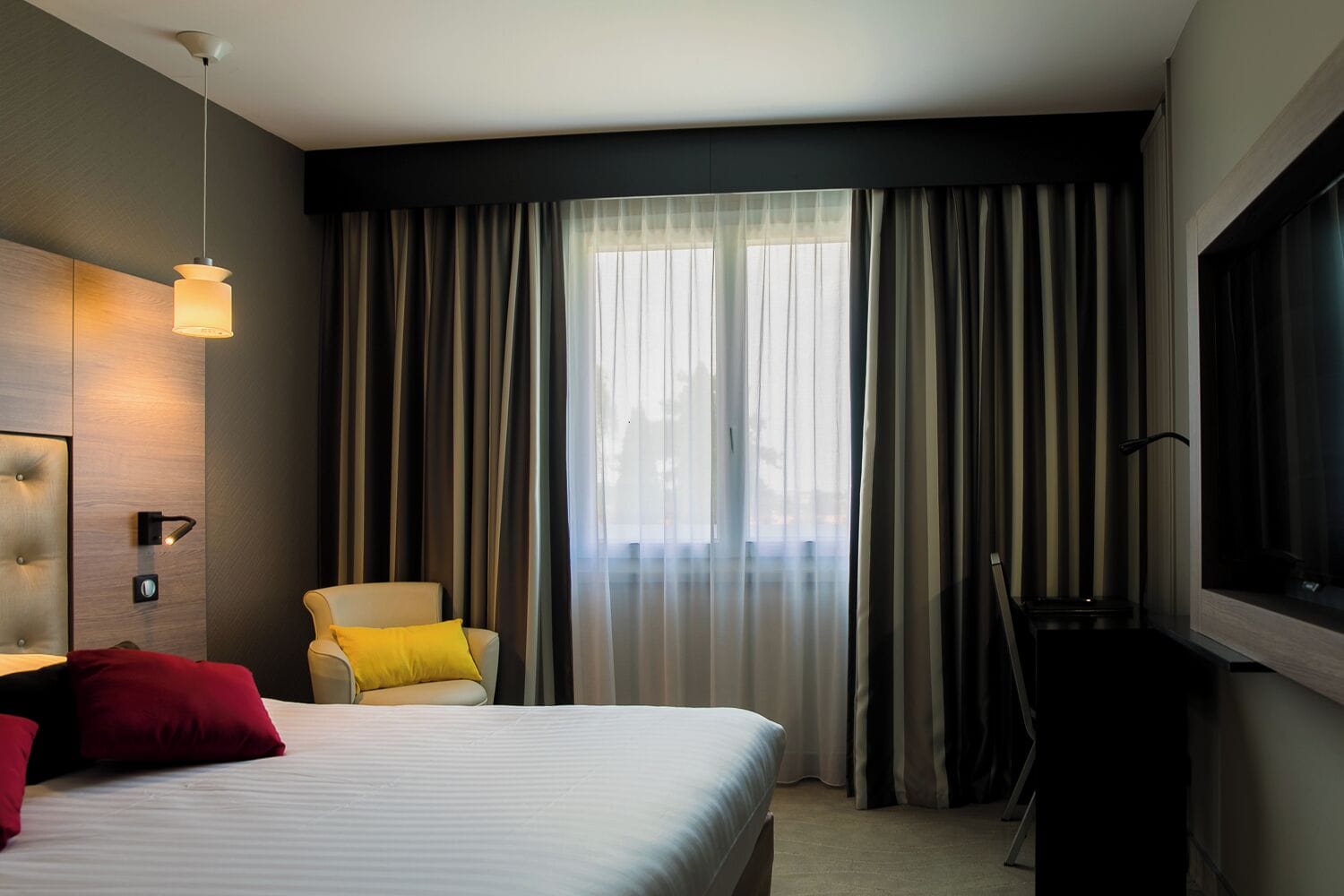 A modern hotel room with a large bed, a chair, a TV, and a window with curtains.