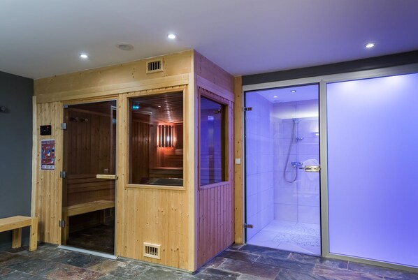 A sauna with wooden walls and a glass door, a shower area with blue-tinted glass, and a stone-tiled floor.