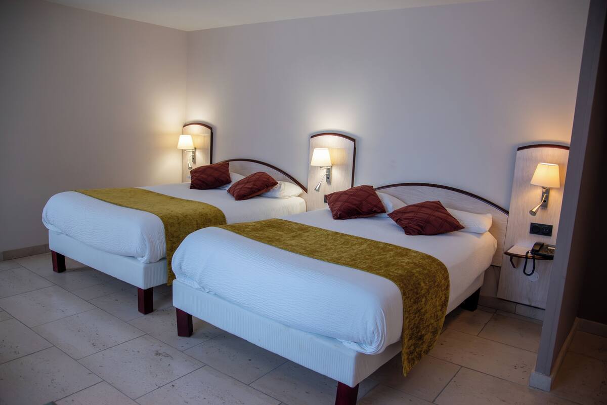 A hotel room with two beds, each with a white mattress and a brown headboard. There are two bedside tables with lamps, and a small desk with a telephone.