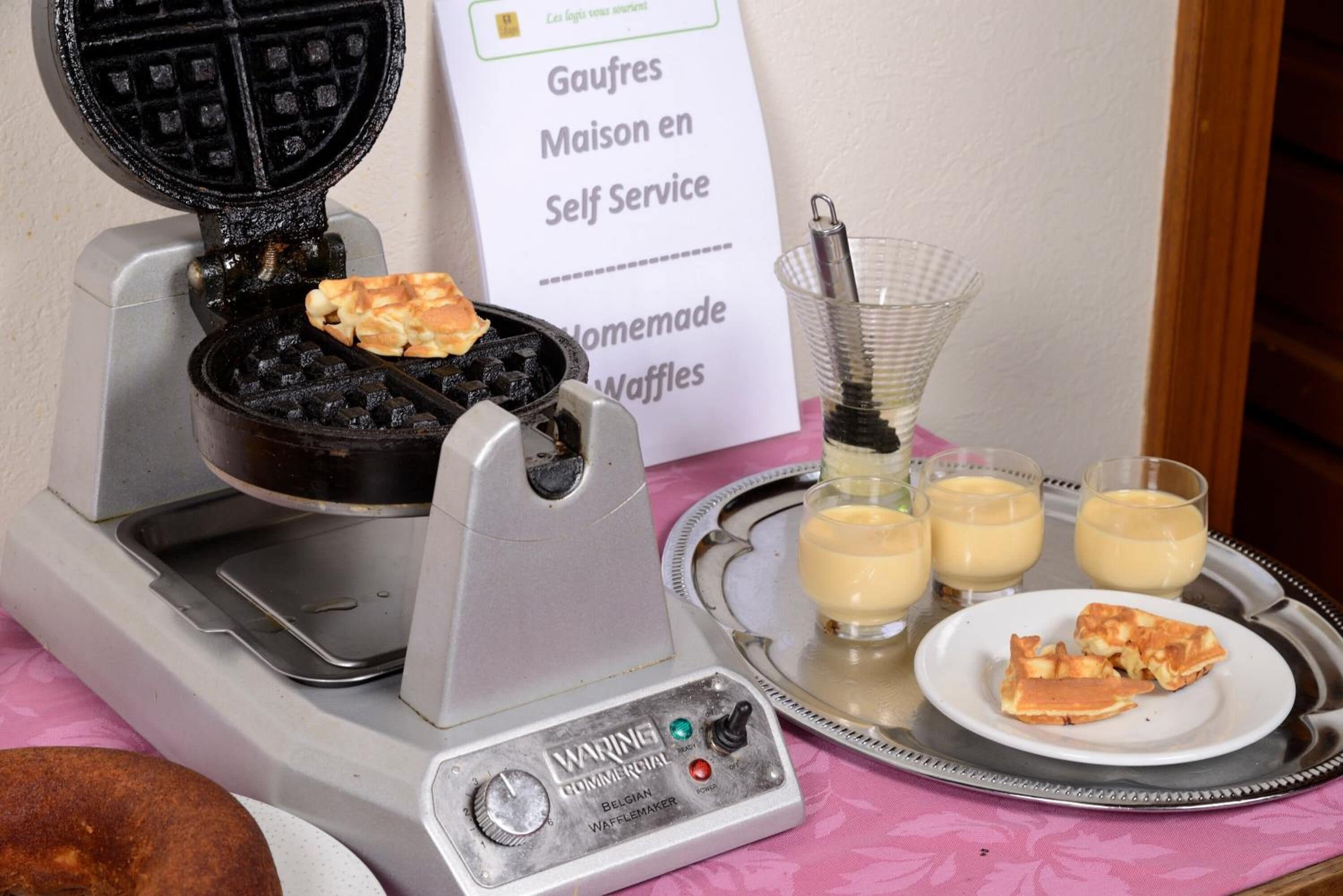 Een wafelijzer met wafels op een dienblad, een bordje voor zelfbediening wafels, en drie glazen met een geel drankje.