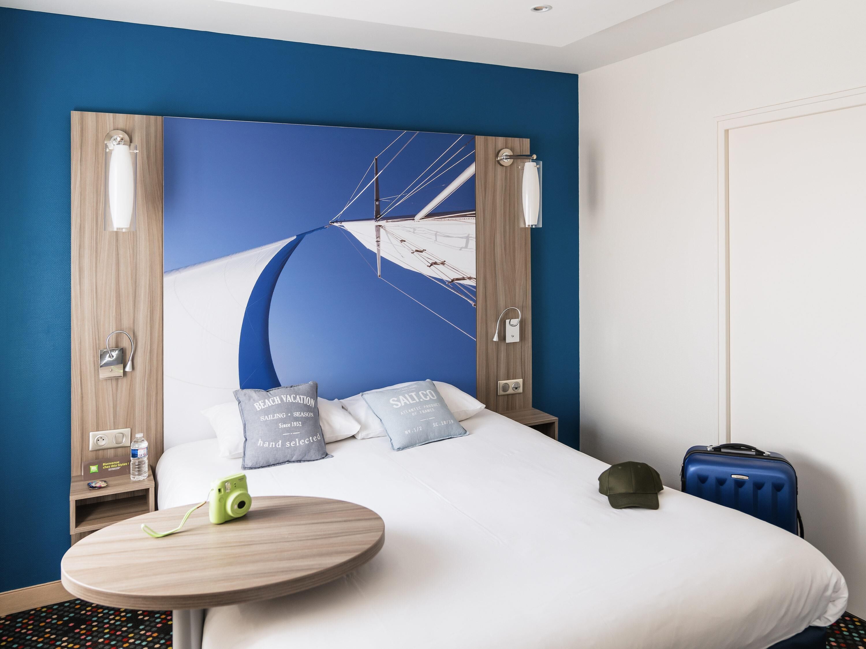 A hotel room with a bed, a round wooden table, a suitcase, and a wall with a blue and white design.
