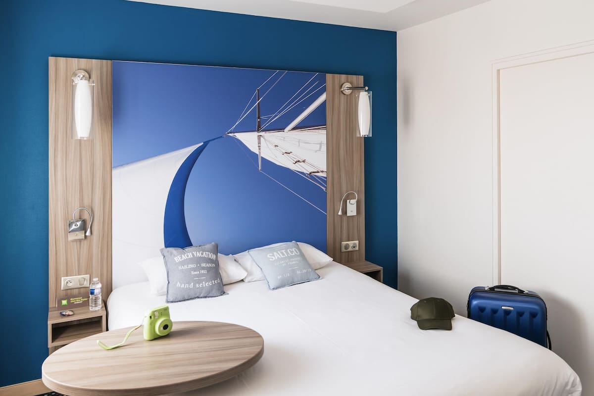 Une chambre d’hôtel avec un lit, une table ronde en bois, une valise et un mur décoré de motifs bleus et blancs.