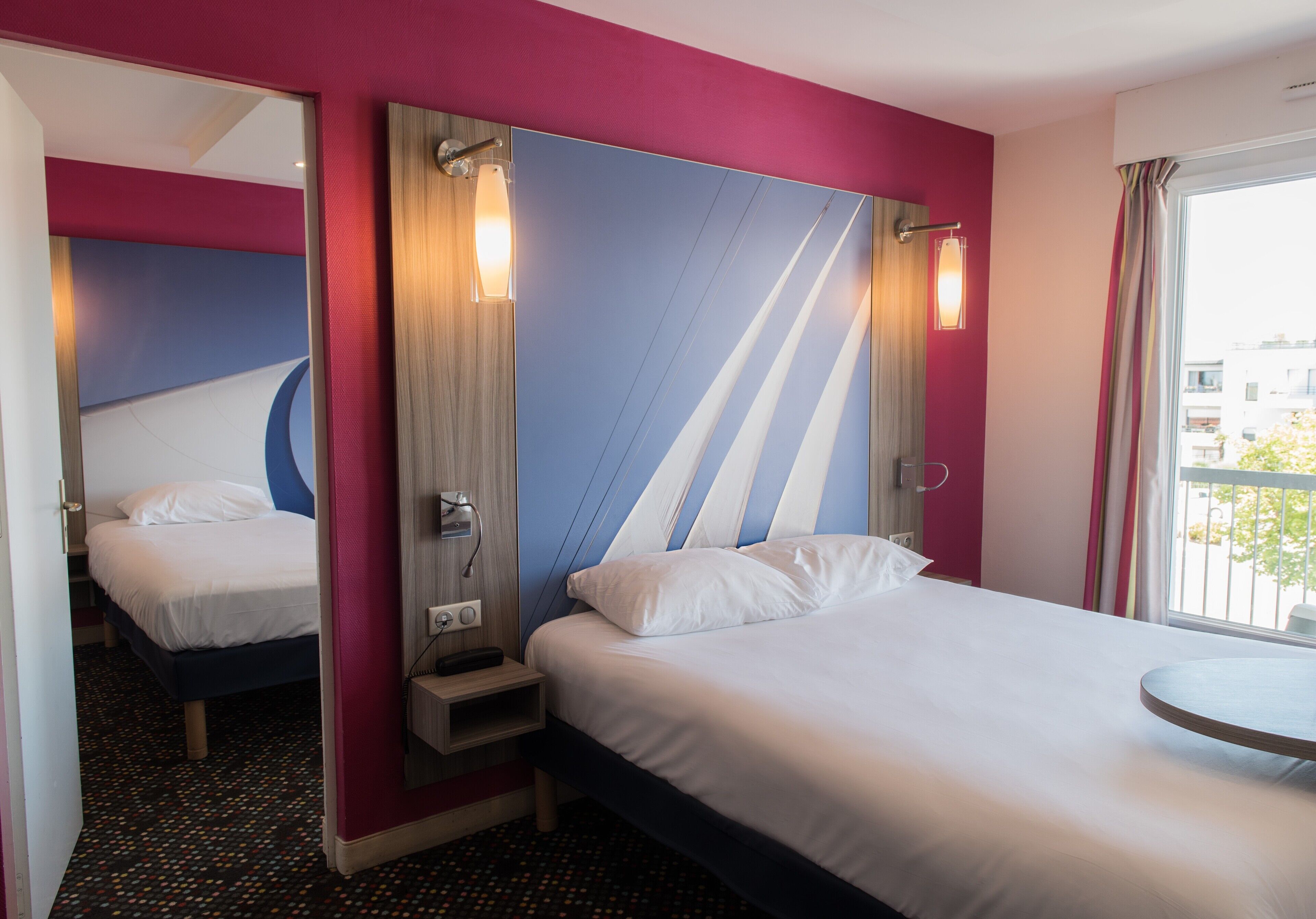 A hotel room with a bed, a bedside table, a mirror, and a window with a view of buildings.